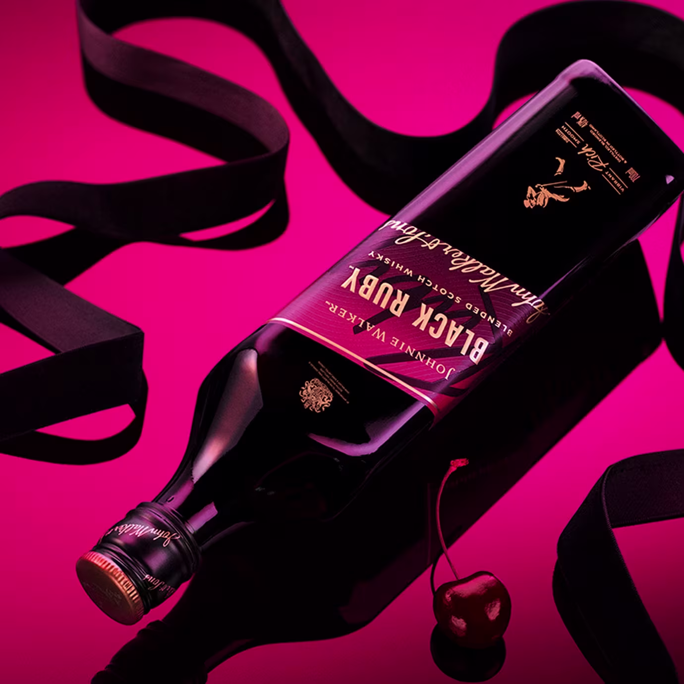 A bottle of Johnnie Walker Black Ruby whiskey lies on a pink background with a cherry and black ribbon nearby.