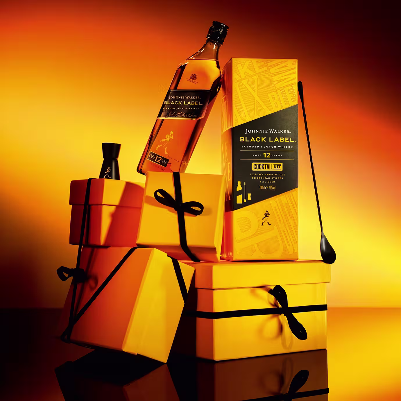 A Johnnie Walker Black Label whiskey bottle and cocktail kit are displayed on stacked yellow gift boxes with black ribbons, alongside a jigger and cocktail spoon, against a warm orange background.