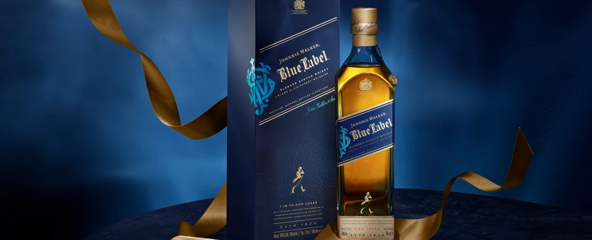 Una botella de whisky Johnnie Walker Blue Label se encuentra junto a su caja azul y dorada, sobre un fondo azul profundo con una cinta dorada envuelta artísticamente a su alrededor.