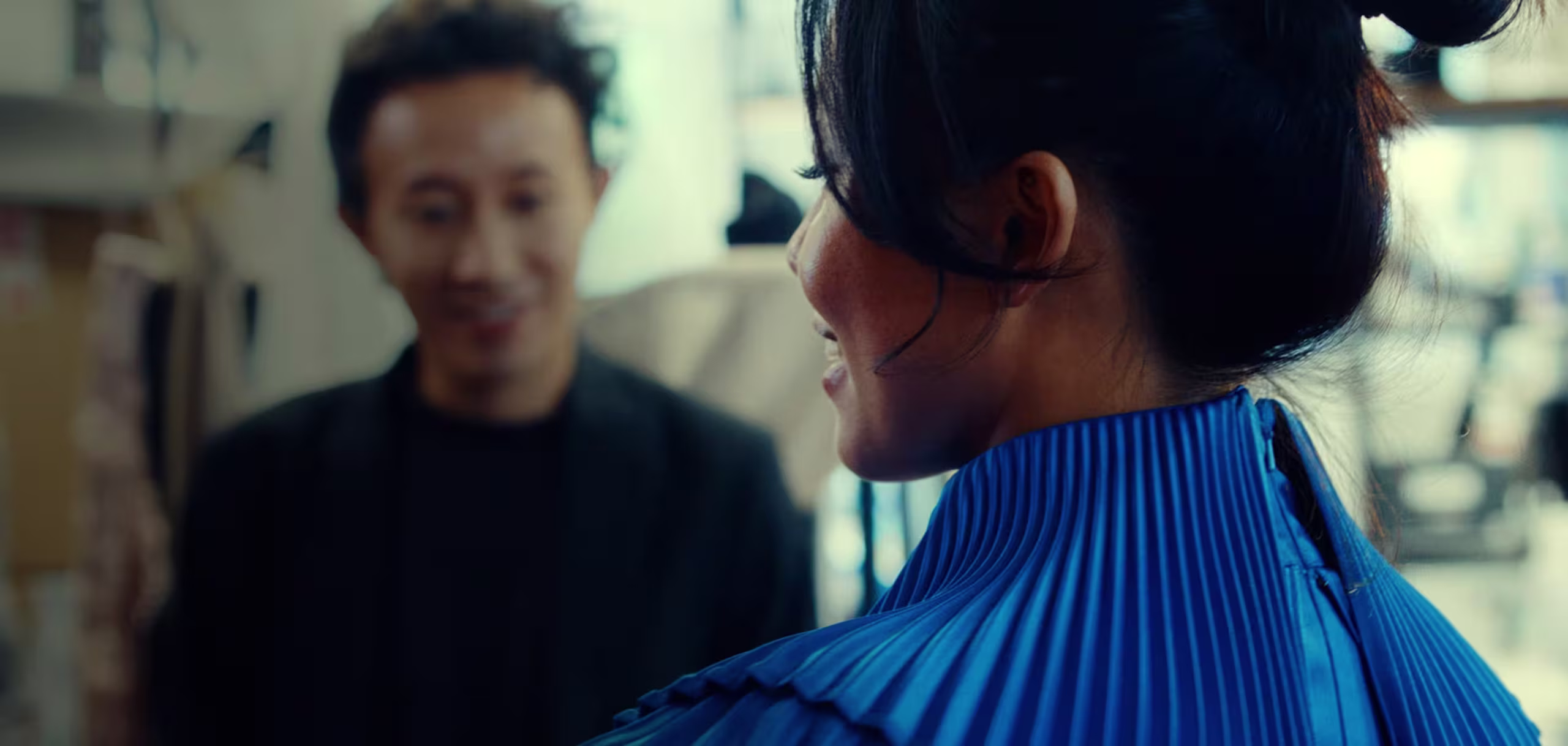 Eine Frau in einem hellblauen, plissierten Outfit wendet den Blick ab und lächelt einen Mann im Hintergrund an, der unscharf ist. Die Szene scheint in einem Innenraum zu spielen, möglicherweise in einem Atelier oder einer Werkstatt.