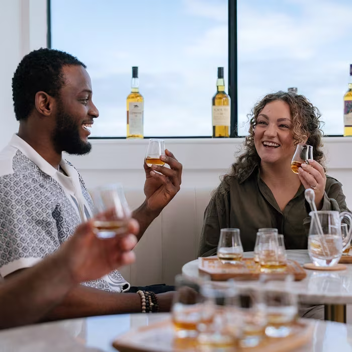 Two people smiling and clinking glasses of whiskey at a tasting, with bottles lined up on the window sill behind them.