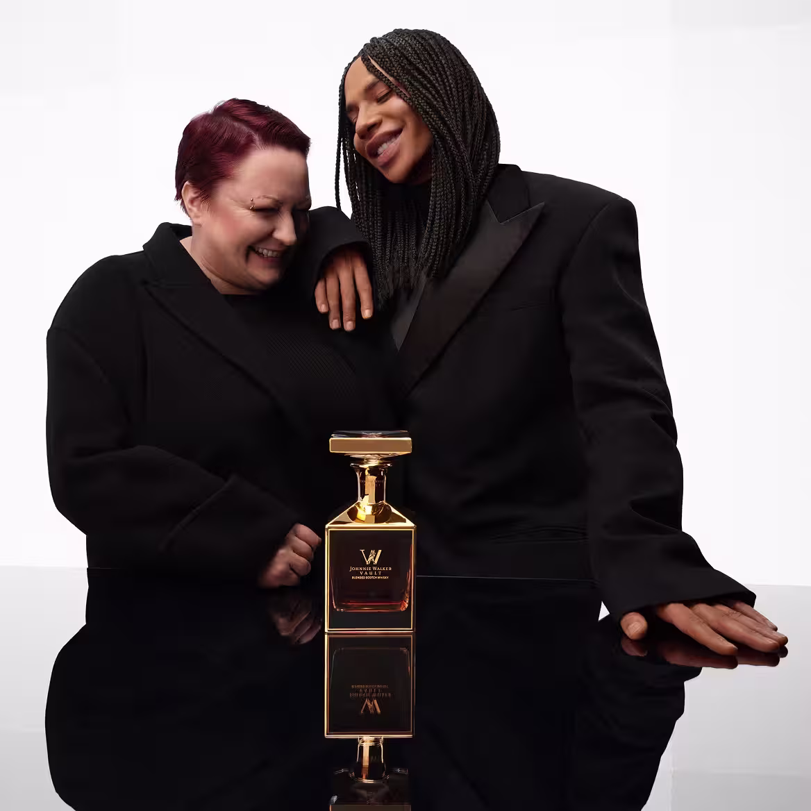 Two people in black outfits smile together behind a shiny, gold perfume bottle on a reflective black surface, against a plain white background.