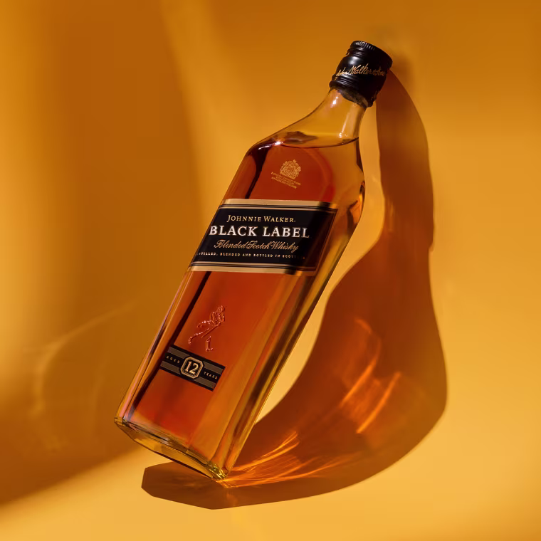 A bottle of Johnnie Walker Black Label 12-year-old blended Scotch whisky lies on its side against a warm yellow background, casting a distinct shadow.
