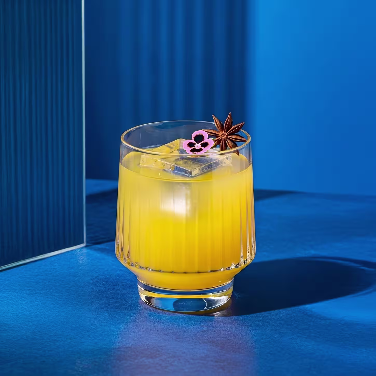 A glass of bright yellow-orange juice with ice cubes, garnished with a pink flower and a star anise, set on a blue surface with a blue background.