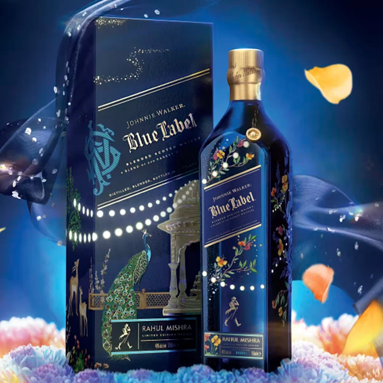 A Johnnie Walker blue label x rahul mishra limited edition design and its decorative box, both featuring intricate, colorful patterns and floral designs. The background includes blue silk fabric, petals, and soft glowing lights for an elegant effect.