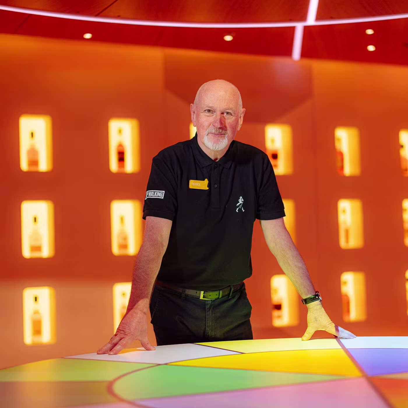 A man in a black polo stands at a colorful table with backlit bottles on an orange wall behind him.