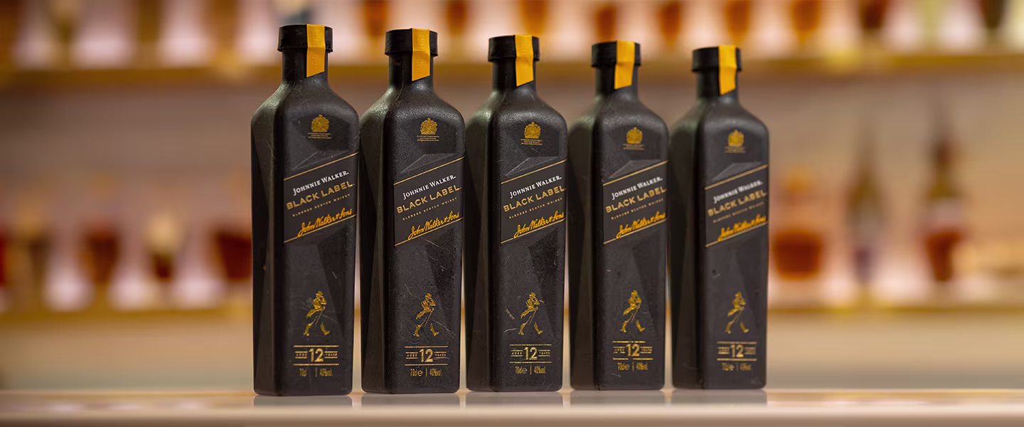 A lineup of five Johnnie Walker Black Label bottles, featuring a first-of-its-kind 70cl paper-based design. The sleek black bottles with gold accents stand against a softly blurred background of whiskey shelves, emphasizing sustainability.