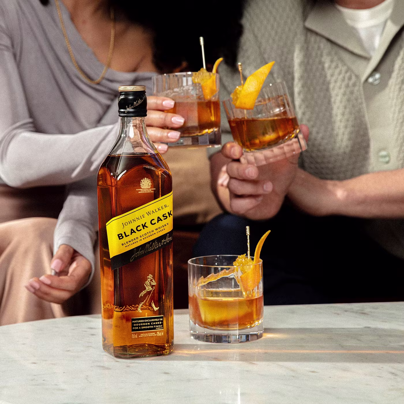 A bottle of Johnnie Walker Black Cask stands on a marble table beside a cocktail glass with an orange twist, while three people toast with similar drinks in the background.