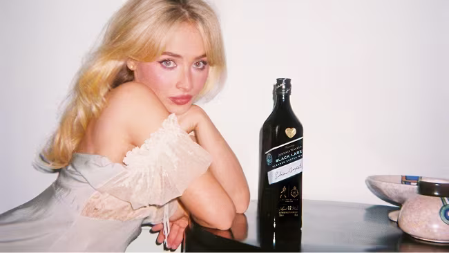 Sabrina Carpenter with blonde hair sits at a table with a dark bottle and dishes in front of a plain white background.