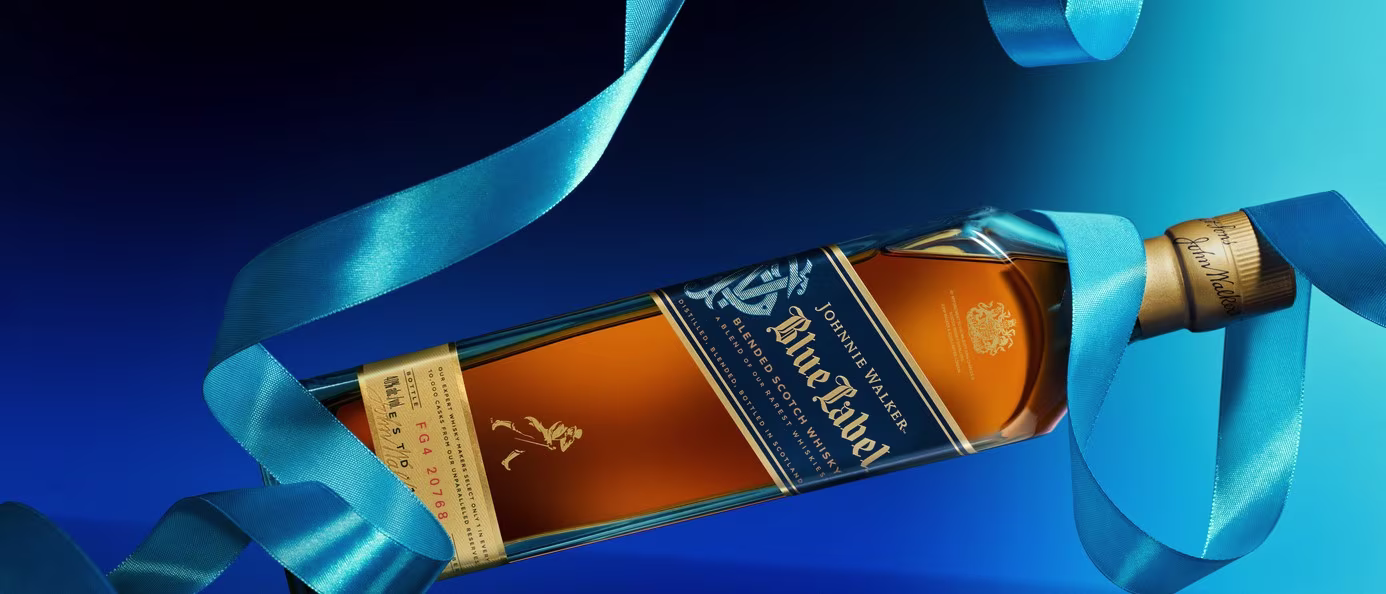 Eine Flasche Johnnie Walker Blue Label Whisky steht auf blauem Grund und ist mit einer glänzenden blauen Schleife umwickelt. Die Flasche ist geneigt und präsentiert so die bernsteinfarbene Flüssigkeit und das elegante Etikett.