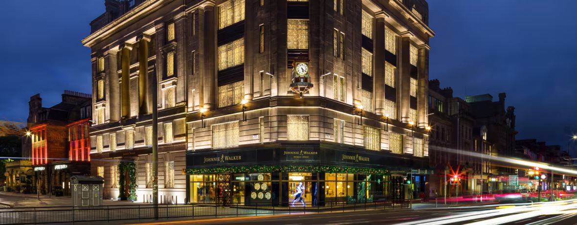 The Johnnie Walker Princes Street building stands tall and proud, its iconic clock tower illuminated against the night sky. The warm glow of the building's lights draws attention to the bustling city street below.