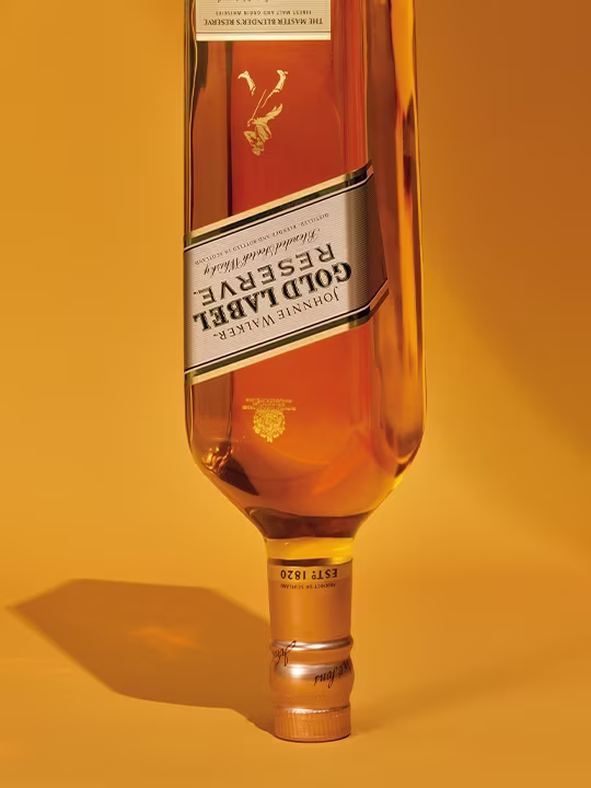 An upside down bottle of johnnie walker gold label reserve whiskey against a warm yellow background. The bottle has a gold and white label with a unique brand logo.
