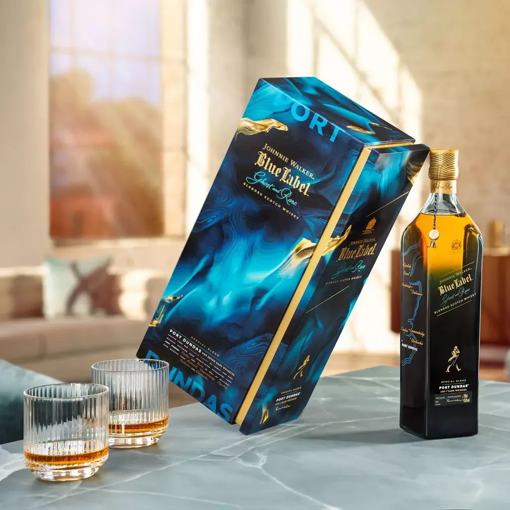 A bottle of Johnnie Walker Blue Label Ghost and Rare whisky on a marble table with two glasses partially filled with whisky. The bottle is next to its blue box packaging. Sunlight streams in through large windows in the background.