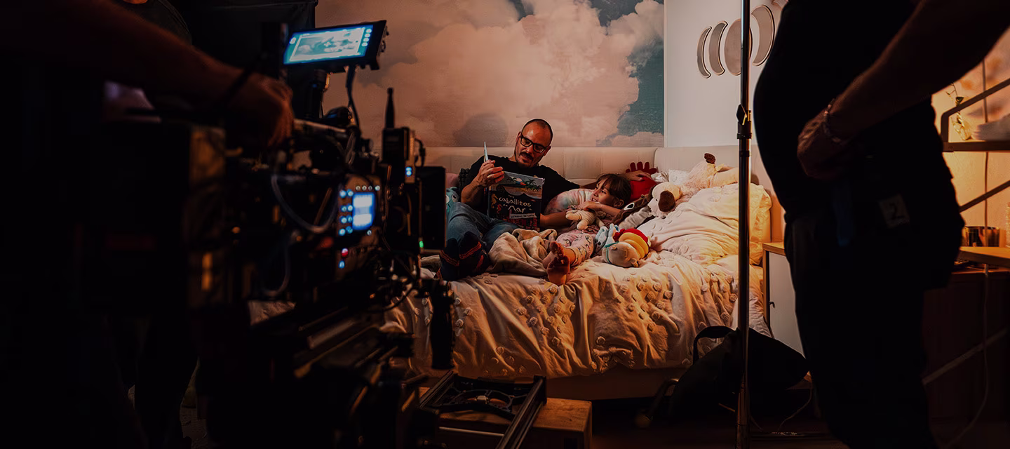 Una escena acogedora en la que un padre lee un cuento a su hijo en la cama, rodeado de peluches. Se ven equipos de filmación y personal, lo que indica que se trata de un plató de cine o de televisión. 