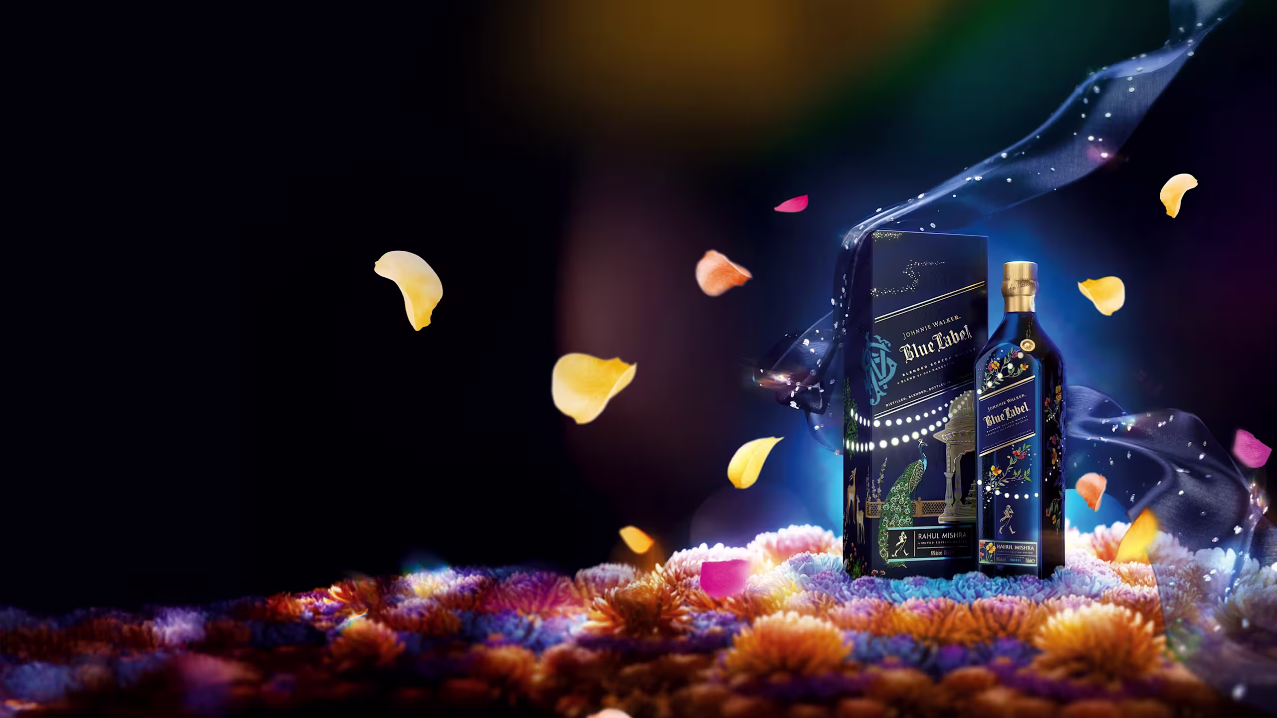 A bottle and box of Johnnie Walker Blue Label X Rahul Mishra sit on a vibrant bed of flowers, surrounded by floating petals and a swirl of blue and purple smoke, set against a dark, dramatic background.