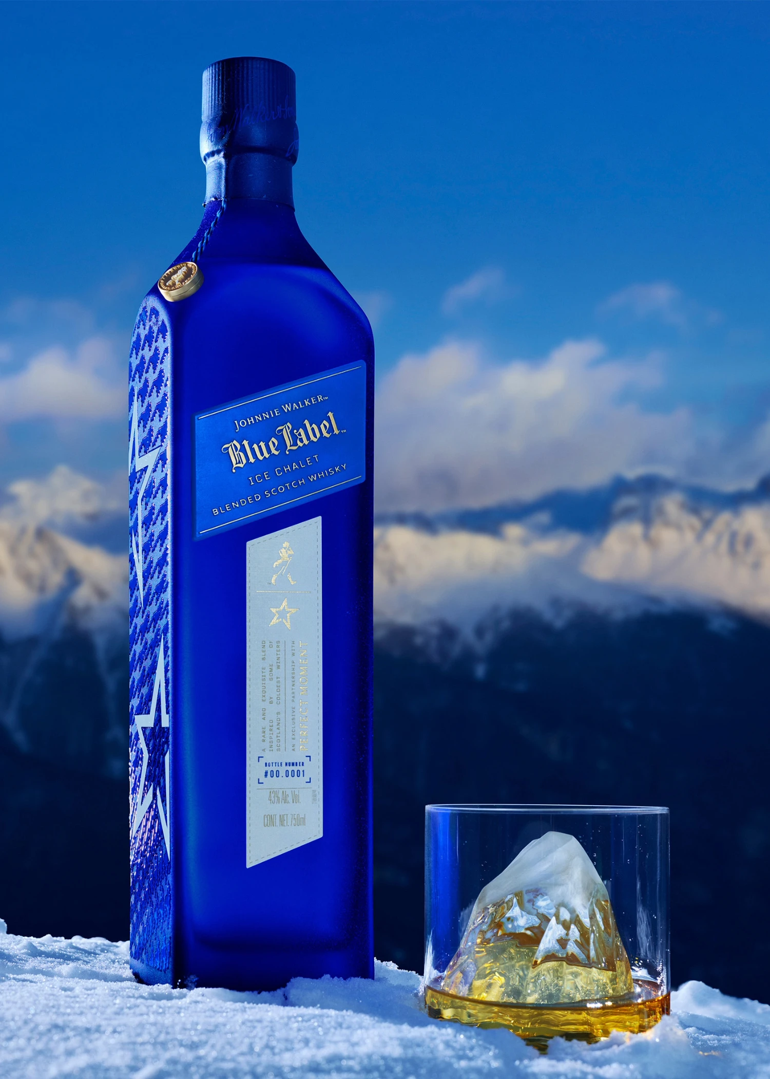 Chalet de hielo con etiqueta azul de Johnnie Walker. Una botella azul de whisky Johnnie Walker Blue Label Ice Edition se encuentra sobre la nieve junto a un vaso de whisky con un cubo de hielo en forma de montaña, con montañas cubiertas de nieve al fondo.