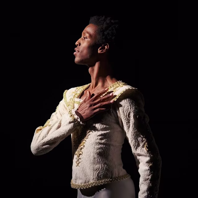 Una bailarina de ballet con un traje blanco adornado con detalles dorados se encuentra de perfil sobre un fondo oscuro, con una mano colocada sobre el pecho, mirando hacia arriba con una expresión elegante.