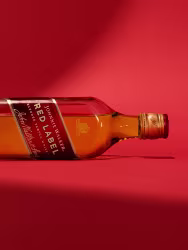 赤い背景にウイスキーのボトルが横たわっています。ラベルには「Red Label」と書かれており、金色のキャップが付いています。照明により、暖かくドラマチックな影が生まれています。.