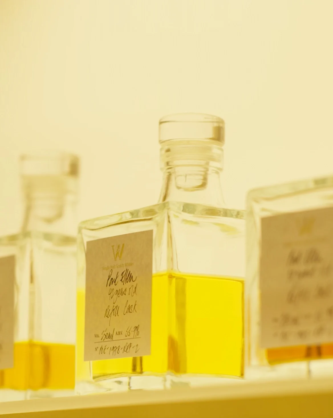 Square glass bottles filled with amber liquid are arranged in a soft, warm light. The bottles have handwritten labels featuring decorative typography.