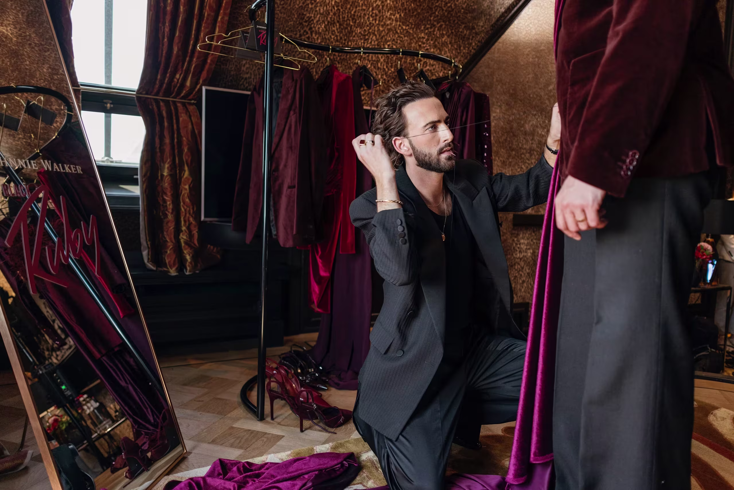 Een modeontwerpster knielt terwijl ze het fluwelen jasje van een staande klant aanpast in een paskamer vol bordeauxrode kledingstukken, schoenen en een grote spiegel.