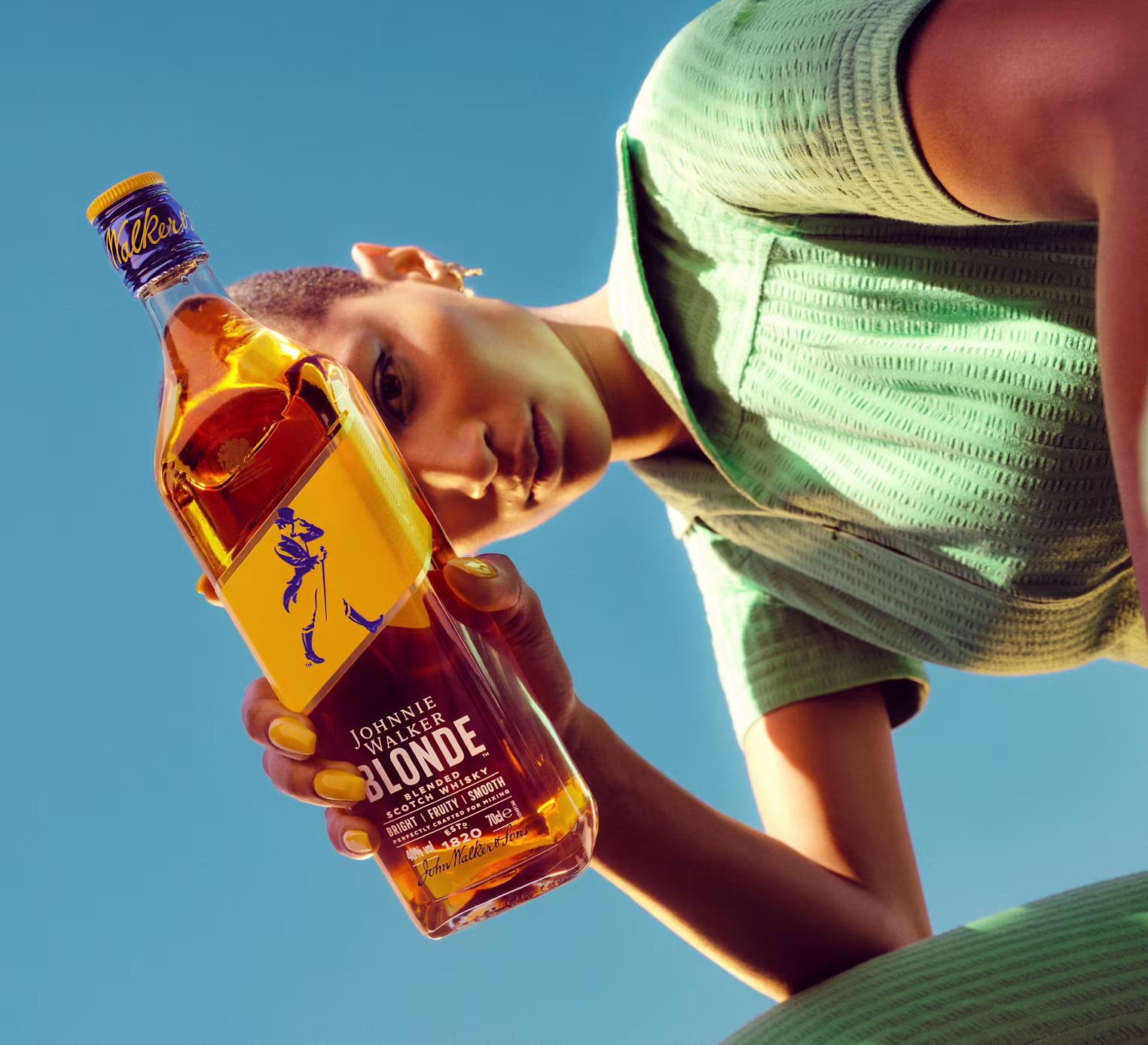 A person in a green outfit holds a bottle of Johnnie Walker Blonde whisky toward the camera against a blue sky, with their face partially visible and looking down.