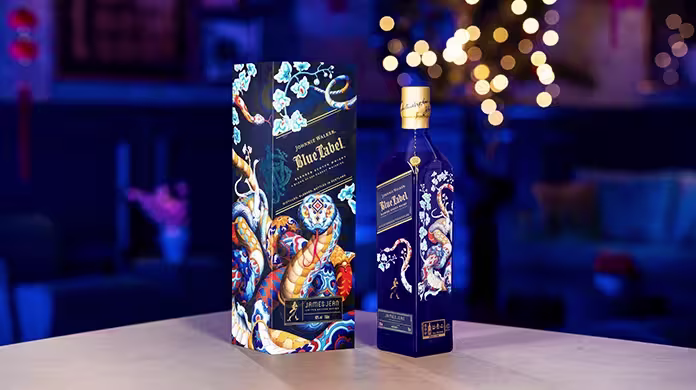 A blue and gold bottle of Johnnie Walker Blue Label stands next to its matching decorative box featuring a colorful dragon and floral artwork, displayed on a table with a blurred, festive background.