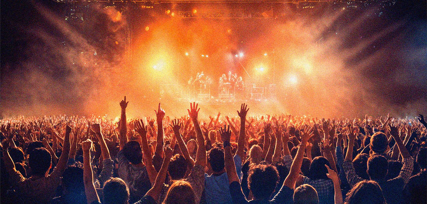 Eine große Menschenmenge mit erhobenen Händen blickt auf eine hell erleuchtete Bühne bei einem lebhaften Konzert oder Musikfestival.
