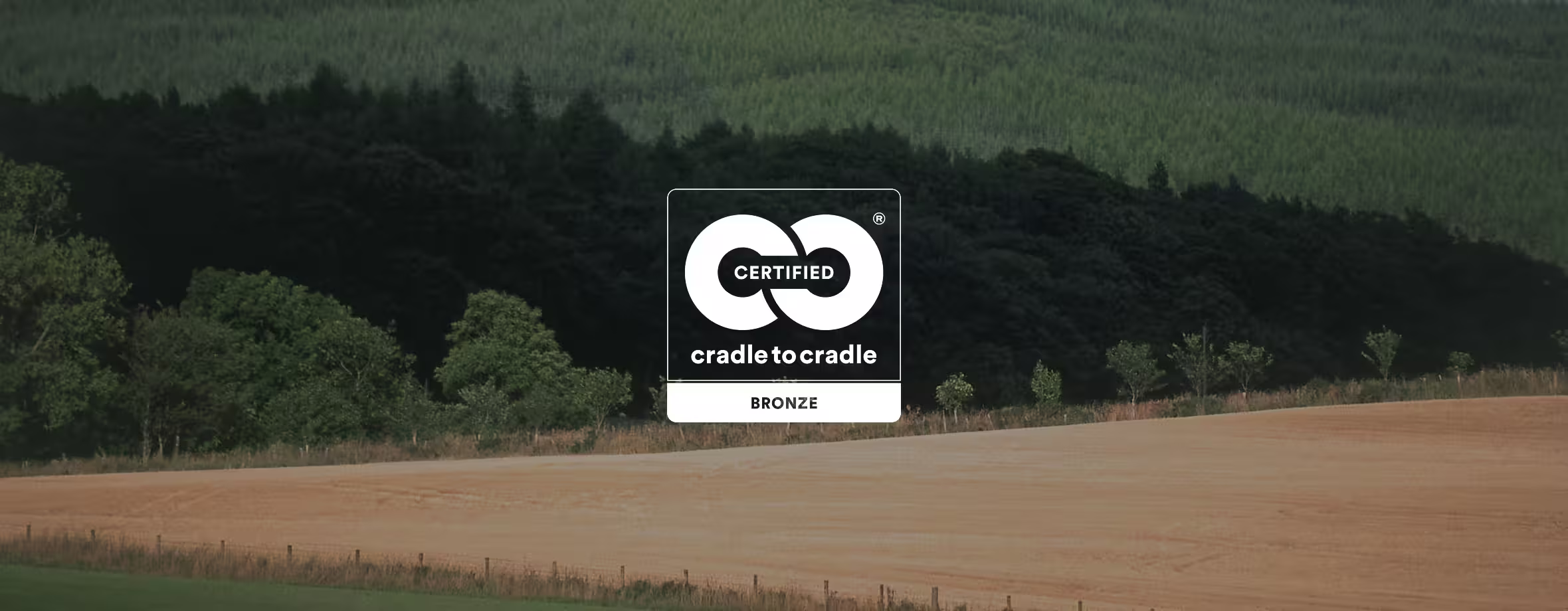 Un paisaje con campos y un bosque al fondo. En el centro, se encuentra el logotipo de Certificación Cradle to Cradle Bronce con dos flechas entrelazadas sobre el texto de certificación.