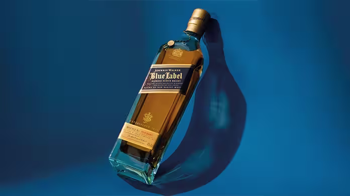 Una botella de whisky Johnnie Walker Blue Label se exhibe de lado sobre un vibrante fondo azul, proyectando una sombra distintiva. La botella tiene una etiqueta azul y un tapón dorado.