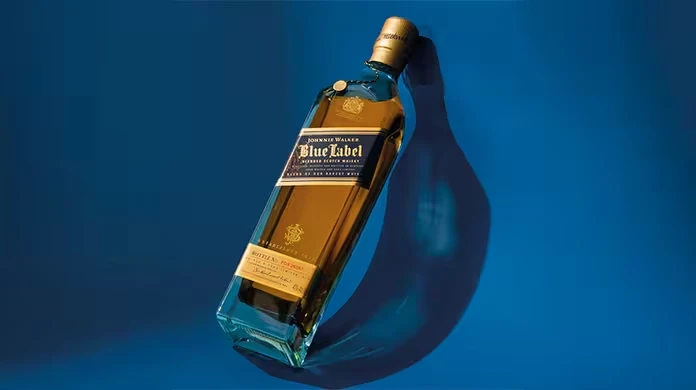 A bottle of Johnnie Walker Blue Label whisky is displayed on its side against a vibrant blue background, casting a distinct shadow. The bottle has a blue label and gold cap.