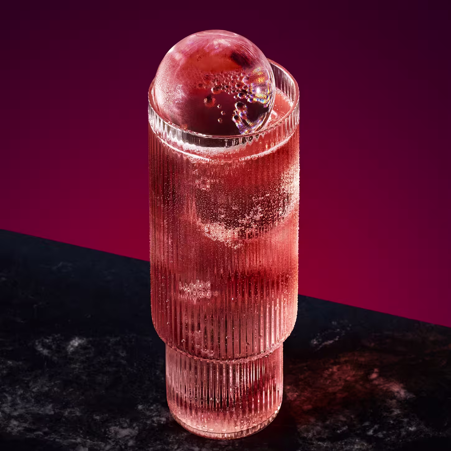 Een hoog, geribbeld glas gevuld met een bruisende roze drank staat op een donker oppervlak. De drank is bedekt met een grote, heldere ijsbol. De achtergrond is een rijke gradiënt van dieprode en paarse tinten.