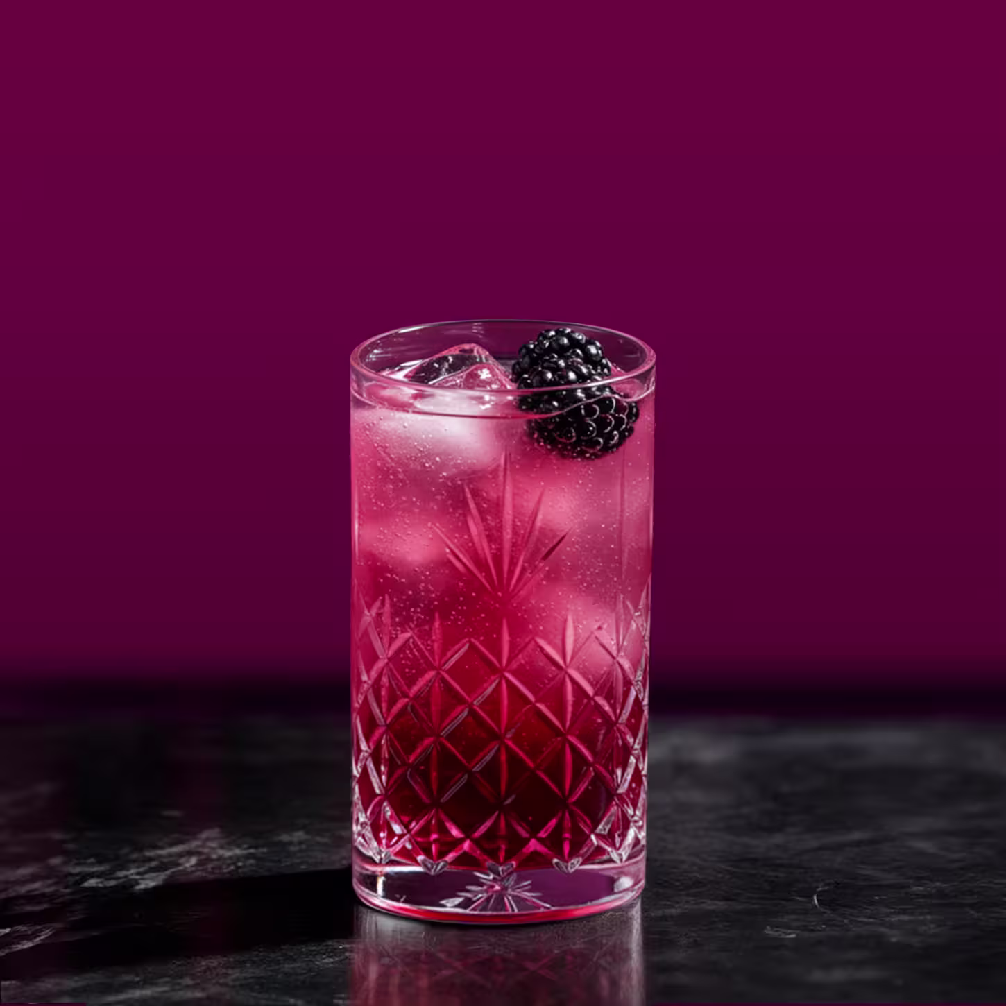 A tall glass filled with a pinkish-purple cocktail, topped with ice cubes and garnished with a blackberry. The glass has a decorative pattern and sits on a dark surface, with a rich burgundy background.