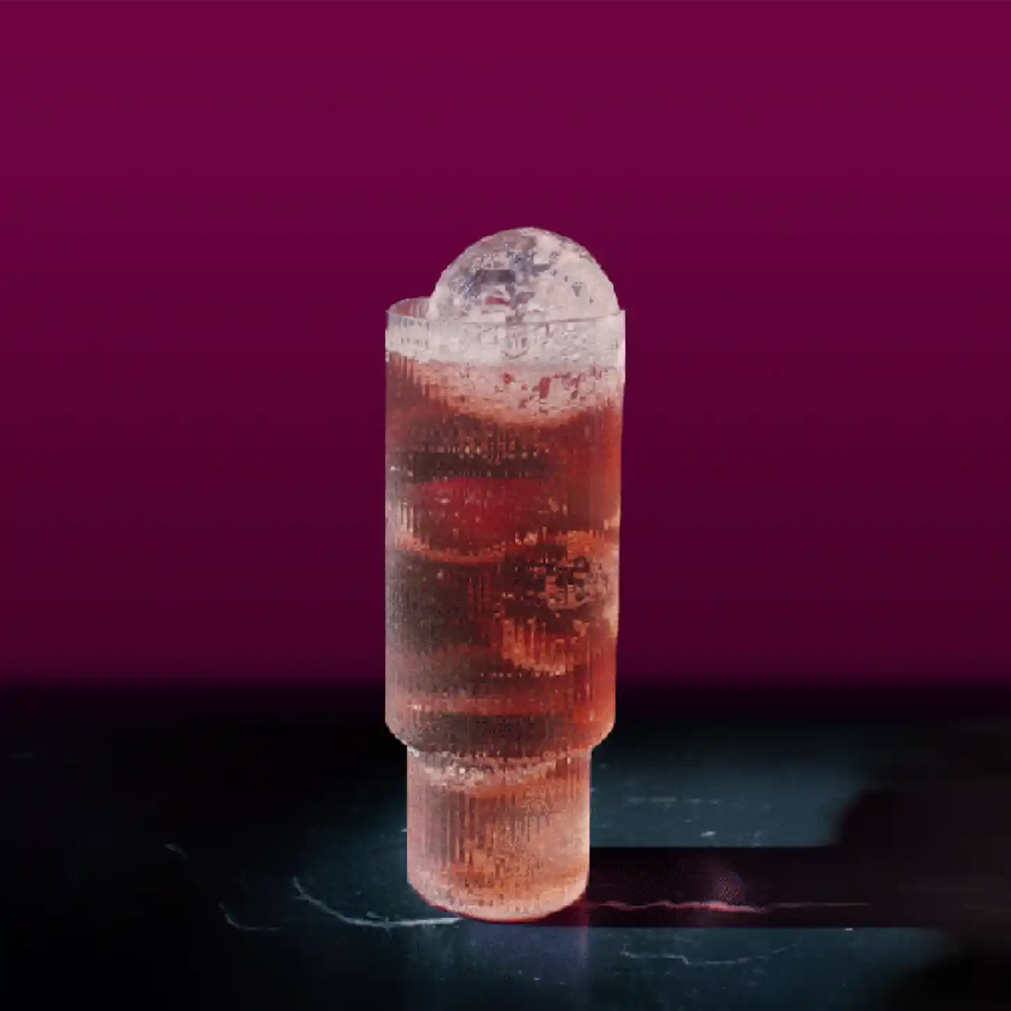 A tall, cylindrical glass with a textured surface holds a reddish-brown liquid topped with a large spherical ice cube. The background is a dark magenta gradient, and the glass is placed on a dark surface.