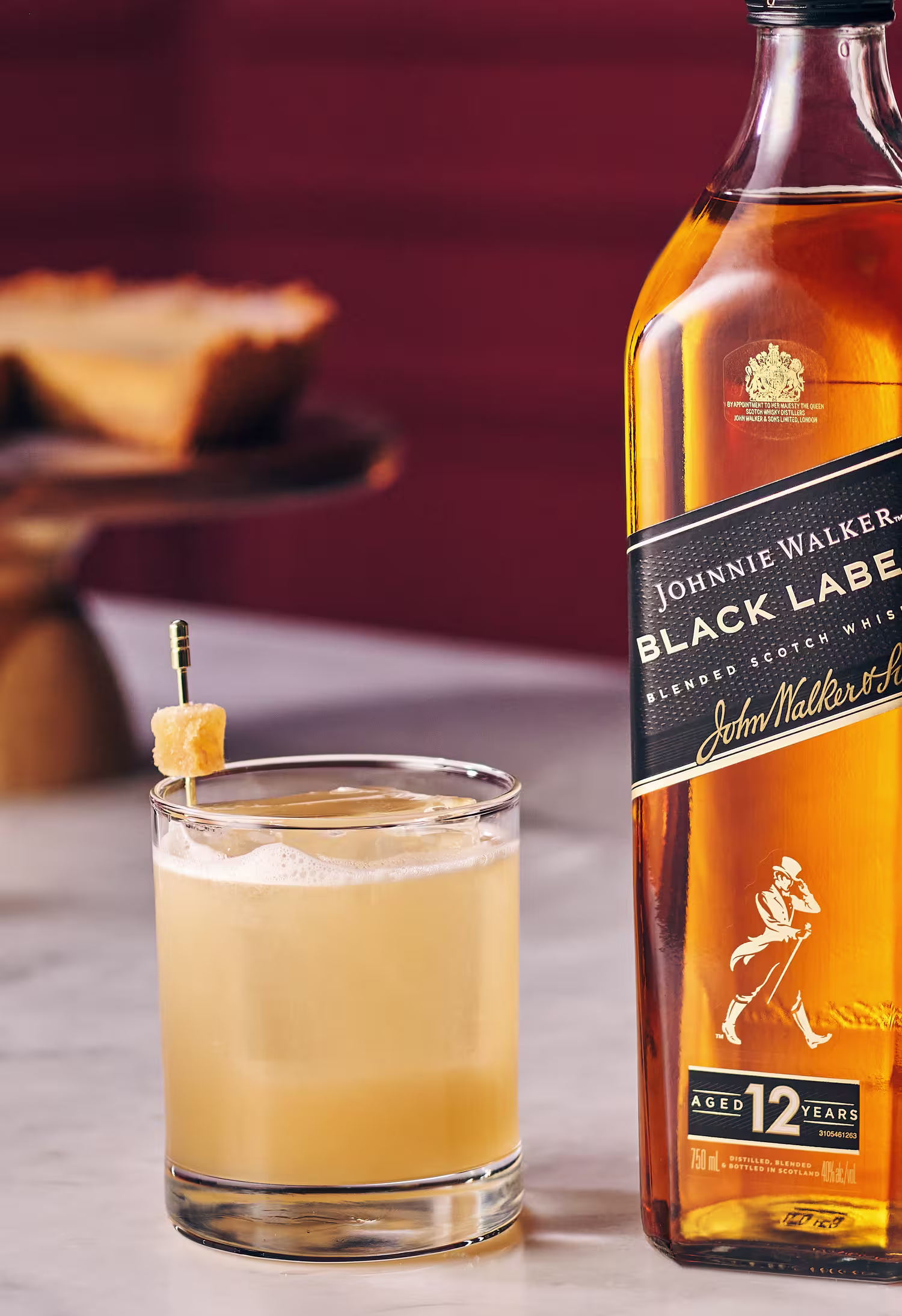A bottle of Johnnie Walker Black Label beside a glass of the Penicilin whisky garnished with a lemon twist on a skewer. A blurred slice of pie is in the background on a wooden stand. The setting has a warm, inviting atmosphere.