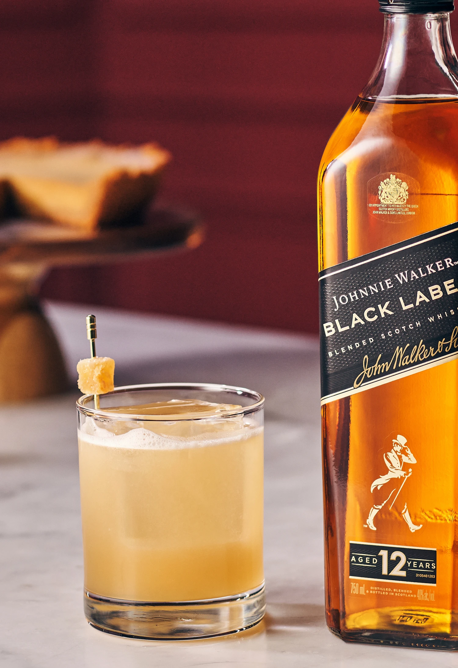 A bottle of Johnnie Walker Black Label beside a glass of the Penicilin whisky garnished with a lemon twist on a skewer. A blurred slice of pie is in the background on a wooden stand. The setting has a warm, inviting atmosphere.