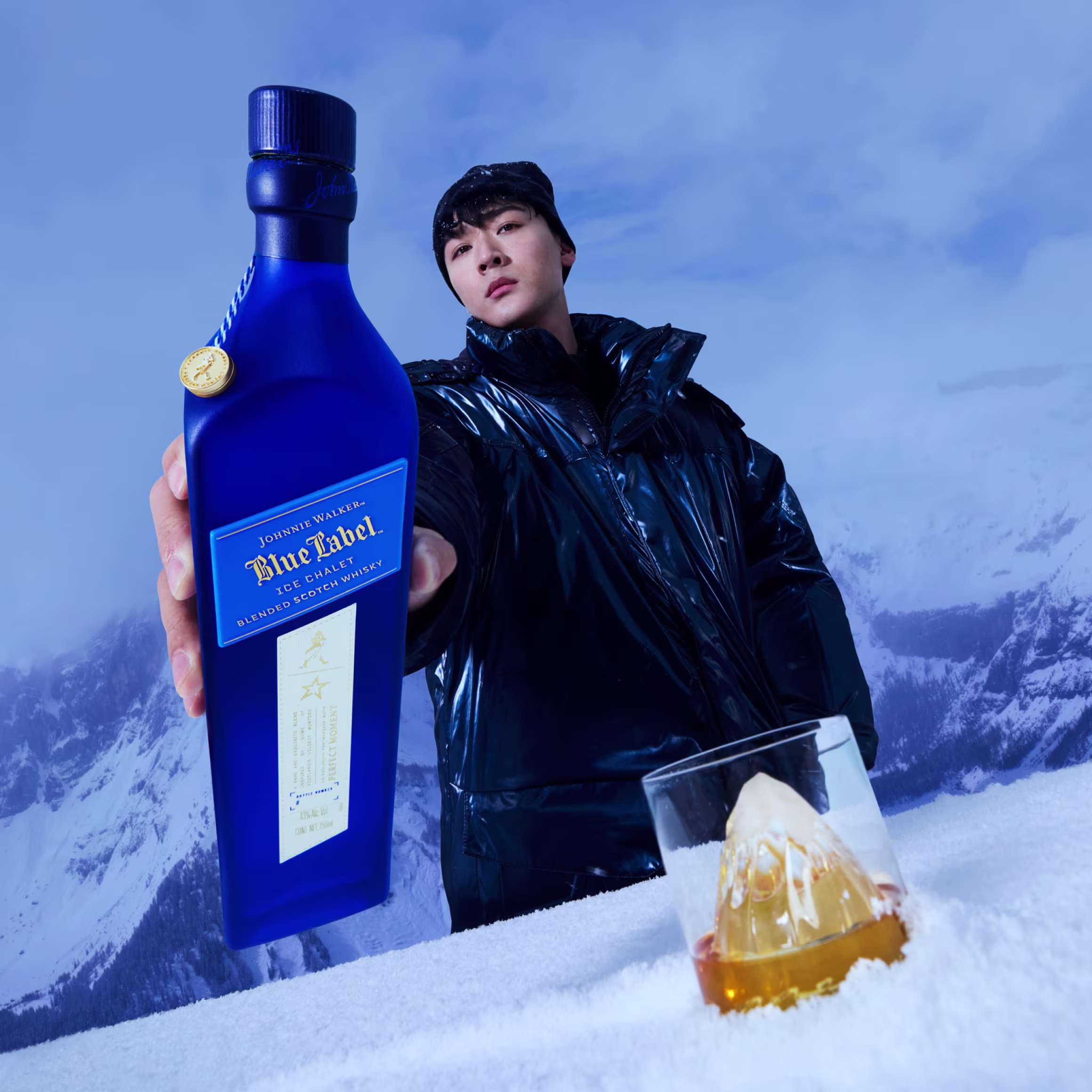 Човек в тъмно палто държи към камерата синя бутилка с надпис „Johnnie Walker Blue Label“. Чаша с кехлибарена течност и кубче лед седи върху сняг. Снежни планини и ясно небе са на заден план.