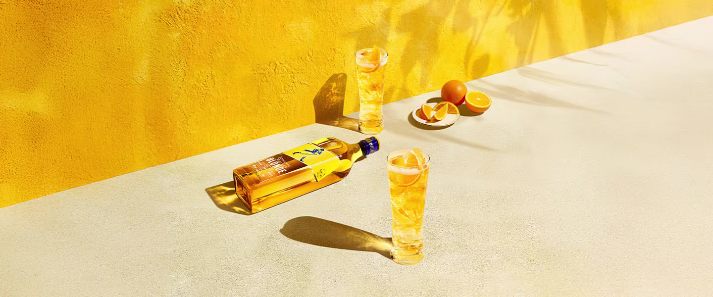 A bottle of amber-colored liquor lies on a light surface against a vibrant yellow wall. Two tall glasses are filled with the same drink. Halved oranges and shadows complete the sunny, refreshing scene.