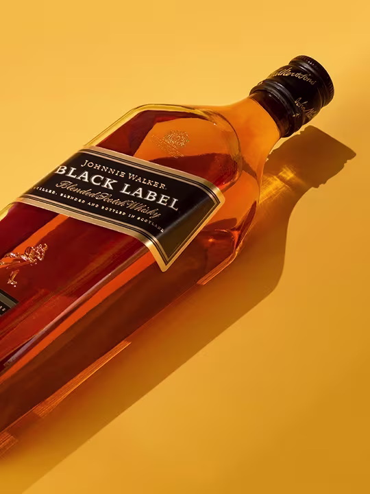 A bottle of Johnnie Walker Black Label Scotch whiskey lies on a yellow surface. The bottle is tilted diagonally, casting a shadow to the right. The amber liquid inside is visible through the transparent glass.