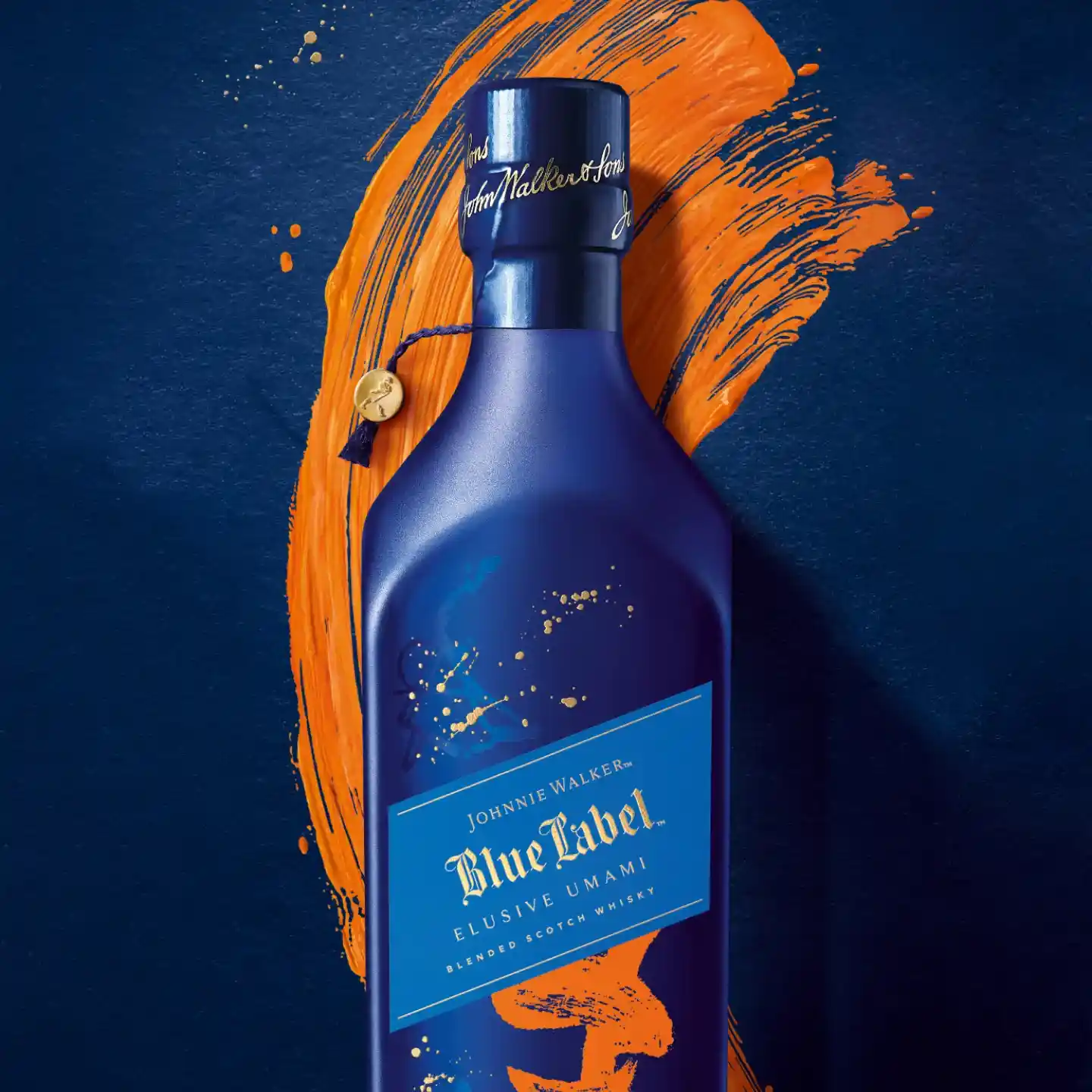 Eine blaue Flasche Johnnie Walker Blue Label Elusive Umami steht vor einem dunkelblauen und orangefarbenen Pinselstrich-Hintergrund. Die Flasche verfügt über eine elegante goldene Beschriftung und ein dekoratives blaues Etikett.