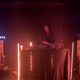 Un DJ con auriculares y una bufanda roja está de pie detrás de una consola, rodeado de barras de luz en una sala con poca luz. Al fondo se ven el equipo y un cartel.