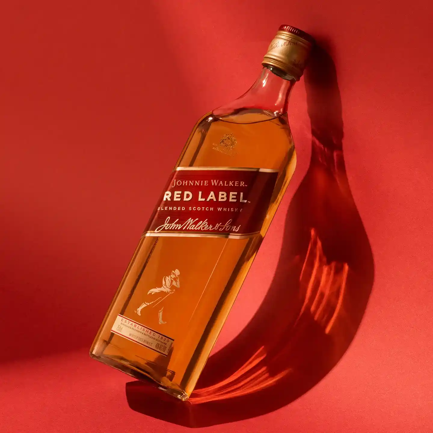 Láhev skotské whisky Johnnie Walker Red Label leží na červeném pozadí. Láhev vrhá stín a její etiketa je viditelná a zobrazuje logo a podpis značky.