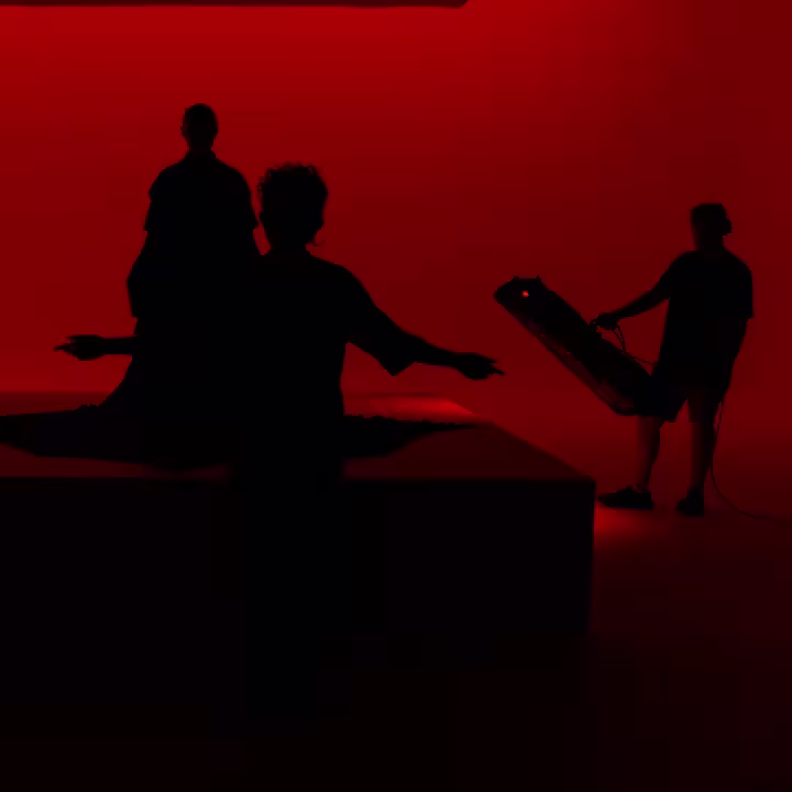 Three people are silhouetted against a red-lit background. One stands on a platform draped in fabric, another stands in front gesturing, and a third person holds a large light panel on the right.