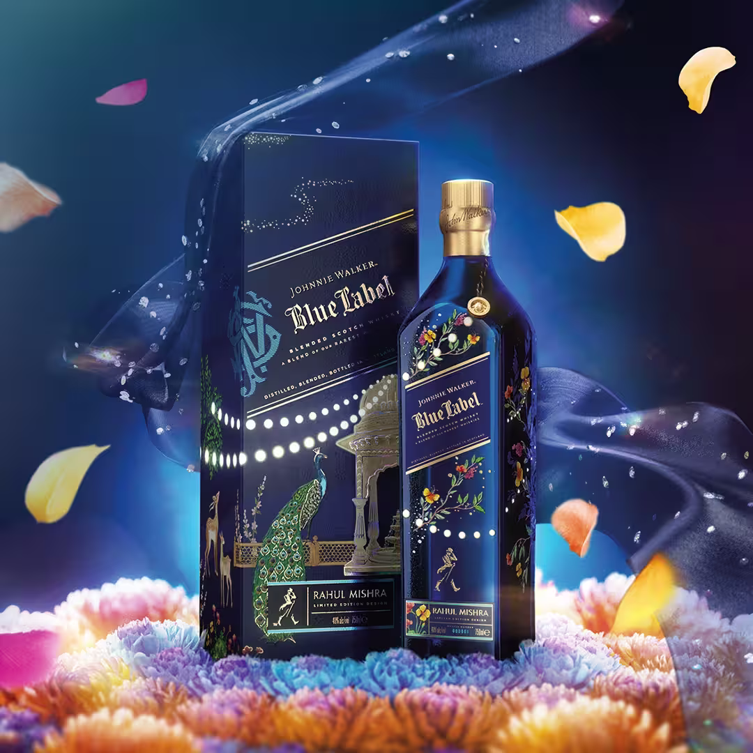 A decorative Johnnie Walker Blue Label bottle and box feature vibrant floral and peacock designs, set against a colourful, festive background with floating flower petals and a starry, dark blue fabric swirl.