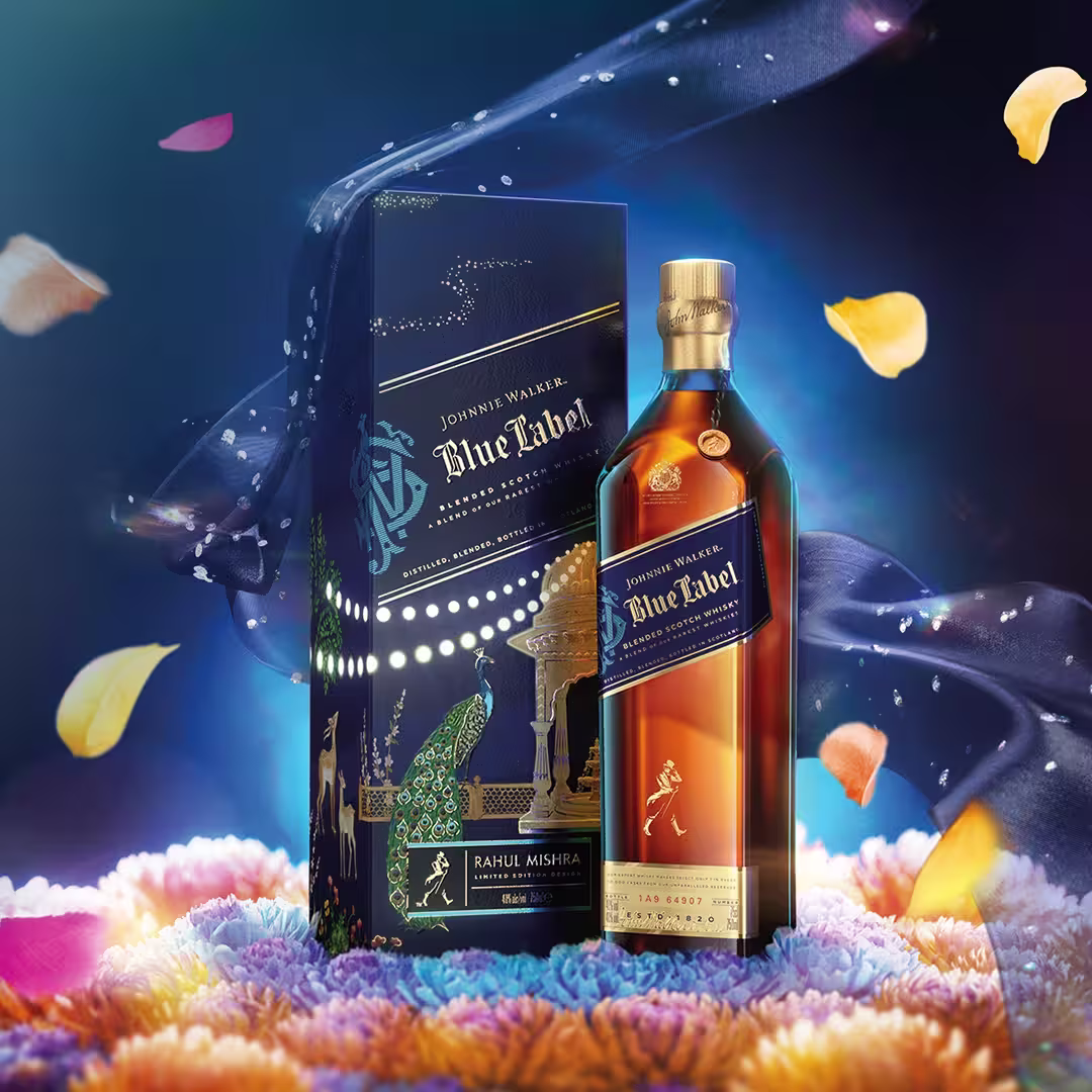 A bottle of Johnnie Walker Blue Label whisky stands beside an ornate, dark blue box with gold and peacock designs. Colorful flower petals float around on a deep, vibrant background.