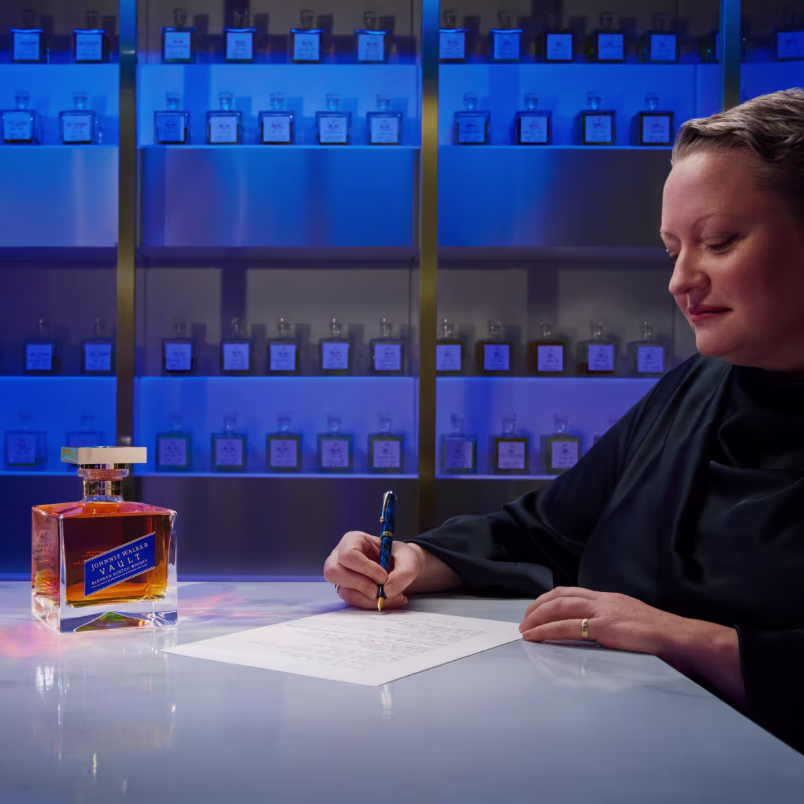 Dr Emma Walker writing notes at a table beside a bottle of Johnnie Walker Vault, with shelves of whisky bottles behind her.