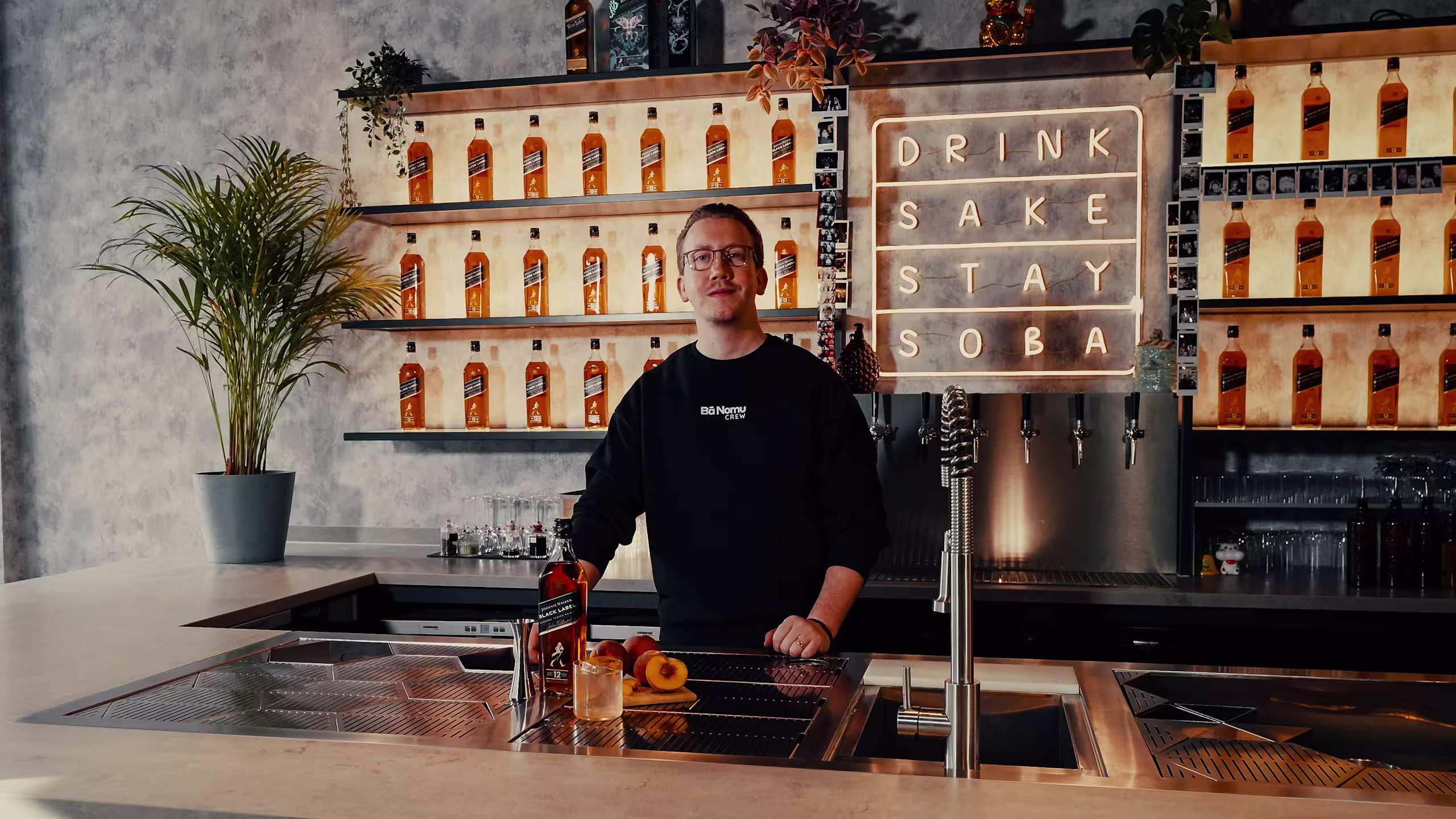 Eine Person steht hinter einer Bar. Im Hintergrund stehen Schnapsflaschen in Regalen. Auf einem Neonschild steht „DRINK SAKE STAY SOBA“. Ein Pfirsich und eine Flasche stehen auf der Theke.