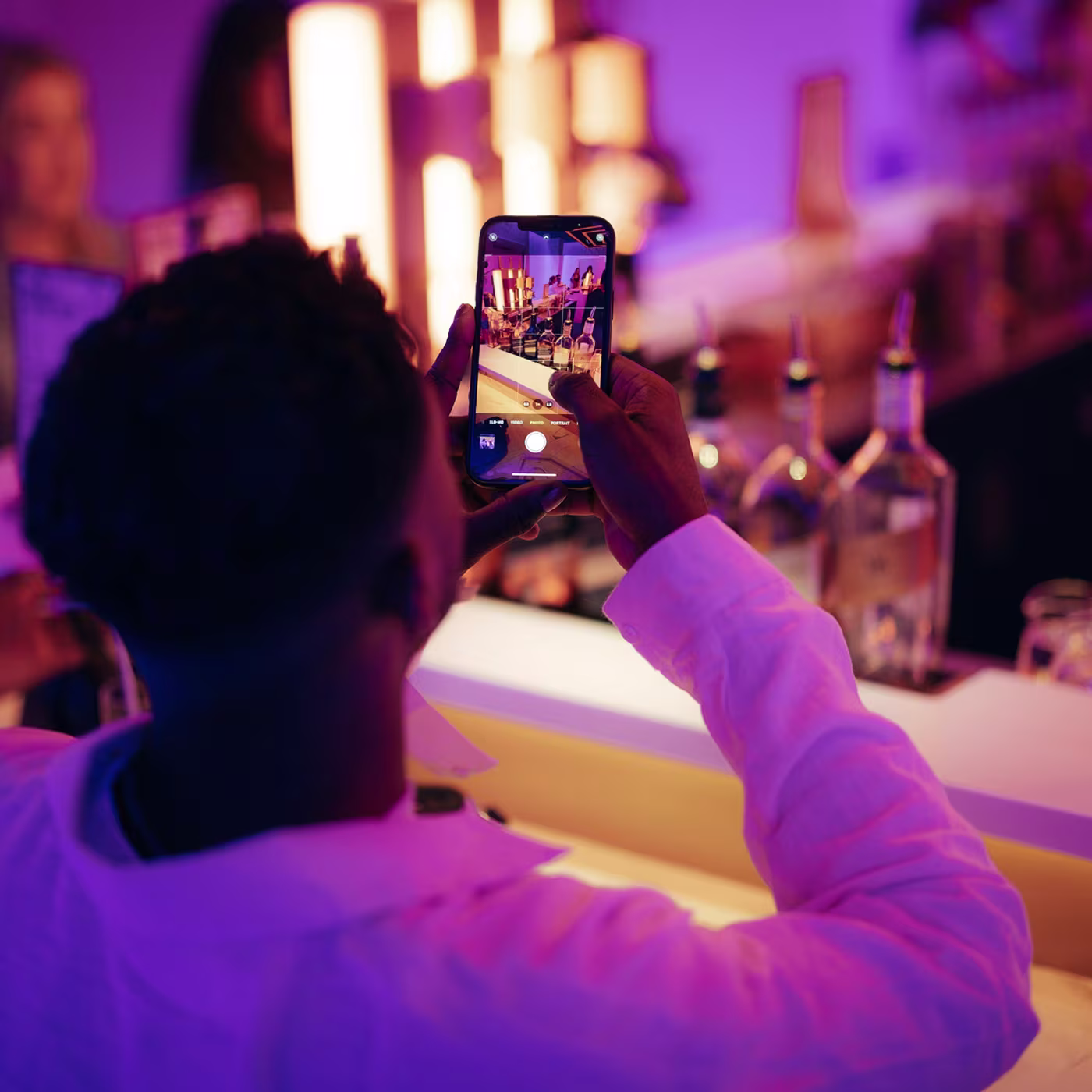 Person taking a photo at a bar with their phone; bottles and neon lights are visible on the counter.