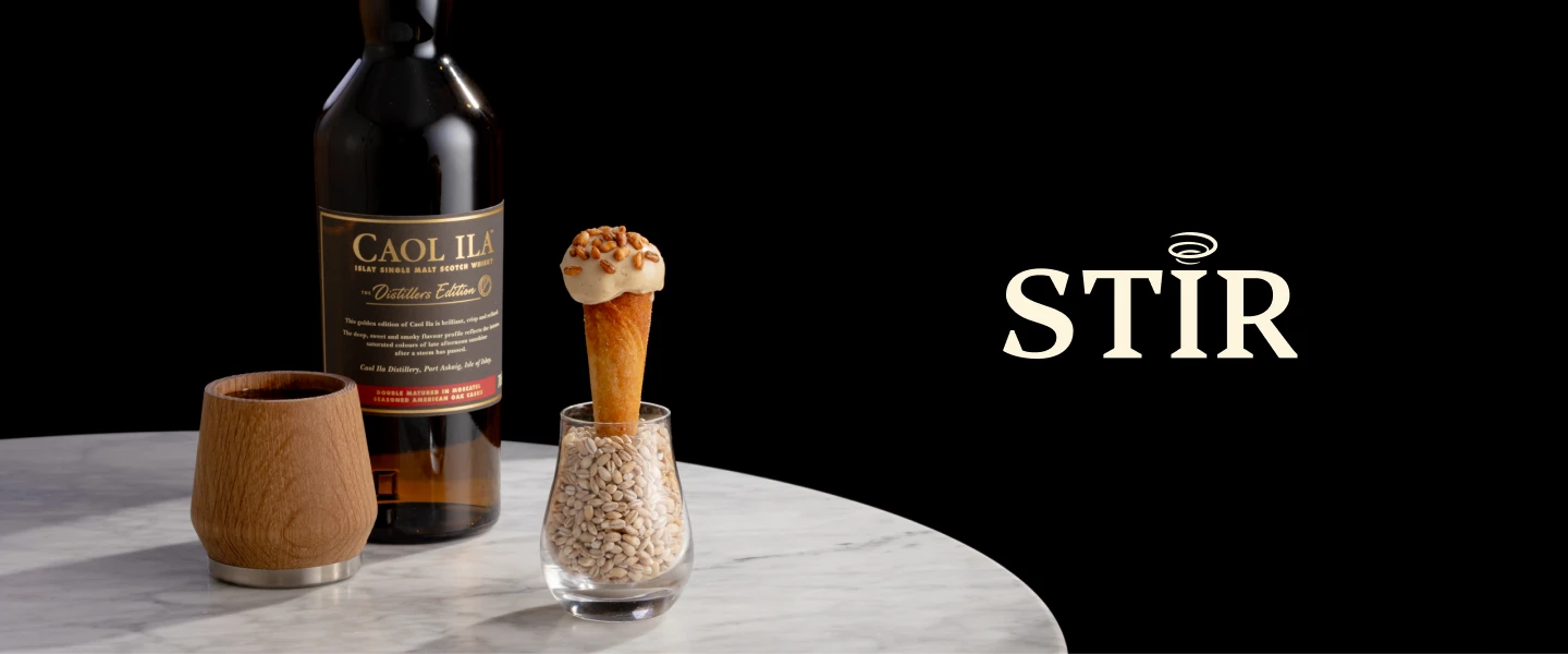 Sobre una mesa de mármol se encuentra una botella de whisky Caol Ila, junto a una taza de madera y un cucurucho de postre lleno de nata y frutos secos en un vaso con granos de cebada. La palabra STIR aparece sobre un fondo negro.
