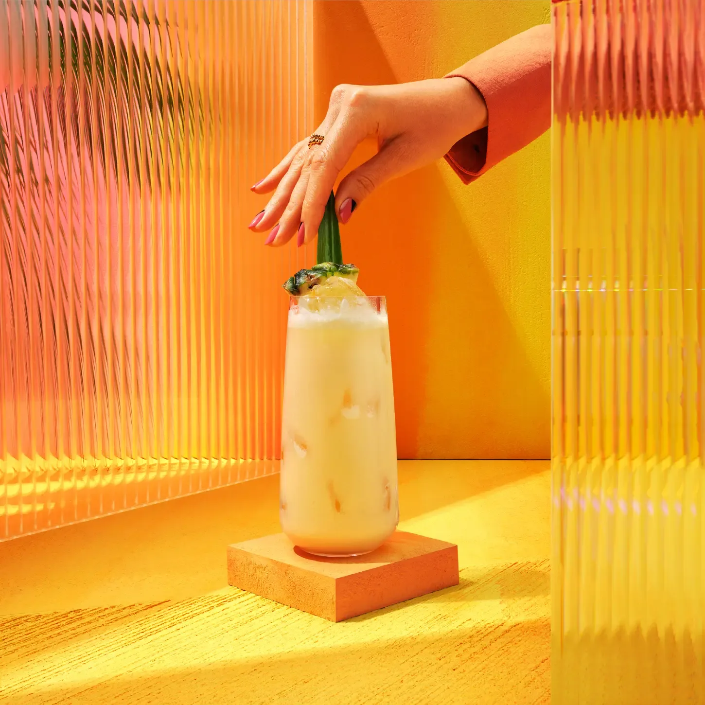 A hand with painted nails gently places a garnished, creamy Pina Colada cocktail on a pedestal. The vibrant setting features orange and yellow hues, with textured glass panels reflecting warm light, creating a tropical ambiance.