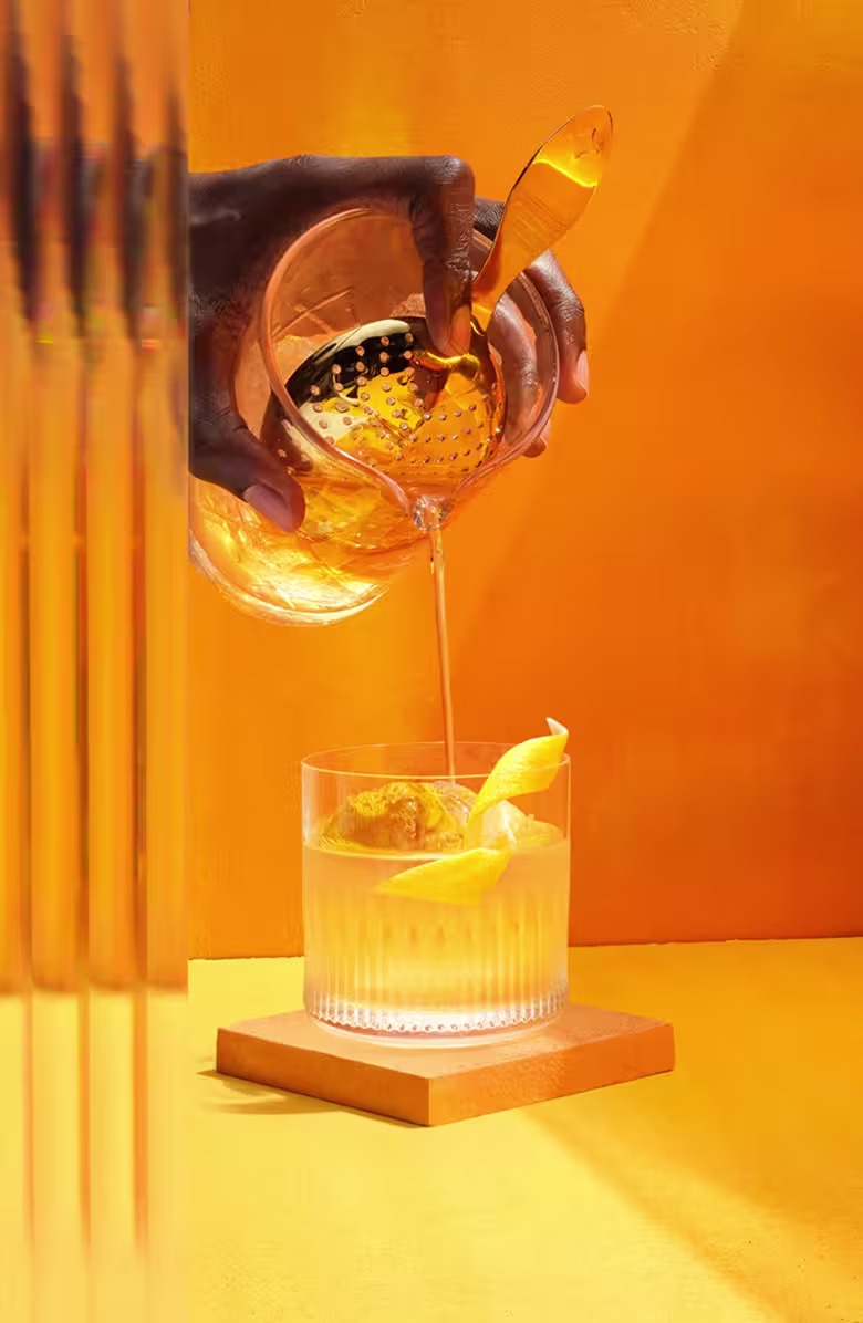 The Smooth & Floral Old Fashioned. A hand with dark skin pours a golden liquid through a strainer into a glass with ice and a lemon twist. The setting is vibrant orange with vertical reflections on the left. The glass is placed on an orange coaster.